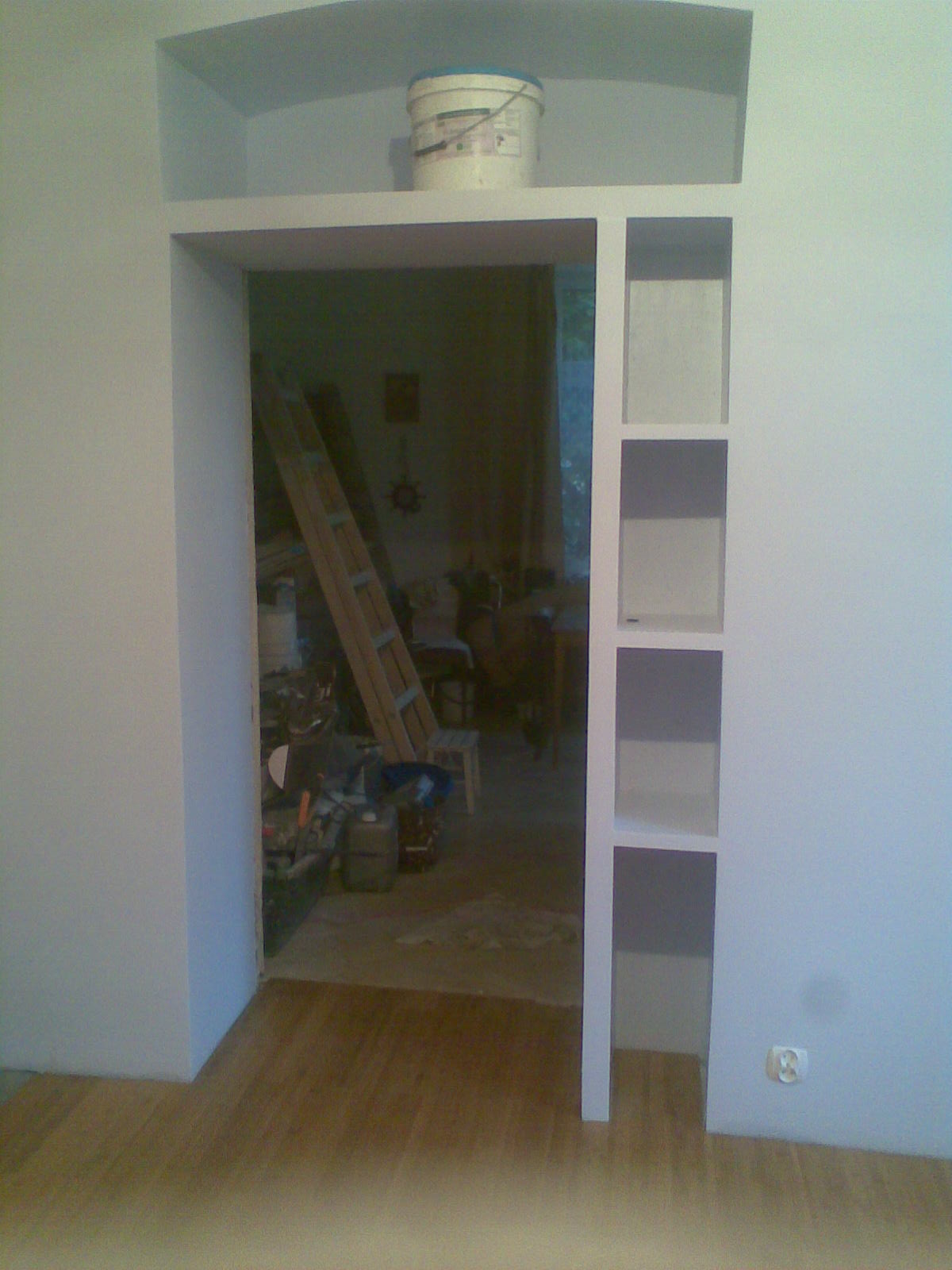 Wnęka w ścianie z półkami, widoczny fragment podłogi i wnętrze pokoju z drabiną i narzędziami, ślady po remoncie.